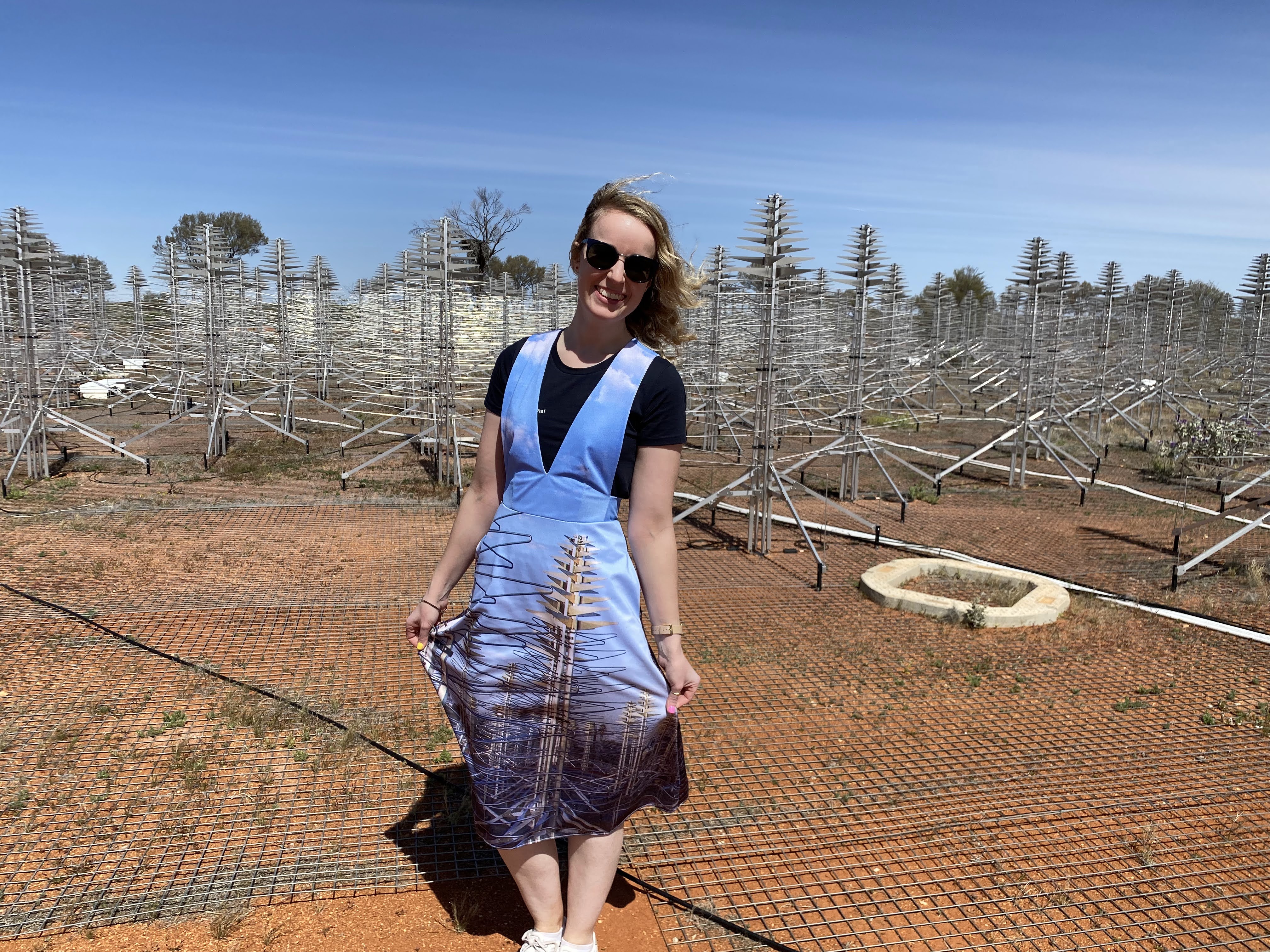 A woman, Dr Laura Driessen, posing in front of a whole bunch of antennas. She has wavy blonde hair and cool glasses with blue rims. She’s wearing a white t-shirt under a pinafore dress. The pinafore dress is light blue and it has two Christmas-tree antennas on it. These antennas are part of the SKA-low test array, so they are antennas for looking at space! The bunch of antennas in the background are the same Christmas tree antennas that you can see on Laura’s dress. This is the SKA-low test array. This is a photo taken by Dr Andrew Zic at Inyarrimanha Ilgari Bundara, the CSIRO Murchison Radio-astronomy Observatory. The sky is a beautiful bright blue with streaky white clouds and the dirt is the lovely orange-red of the Australian outback.